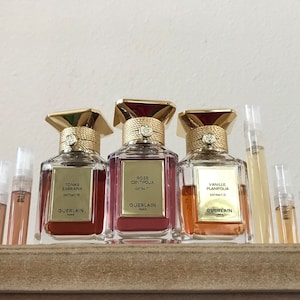 May include: Three glass bottles of perfume with gold caps and labels. The bottles are arranged in a row on a wooden shelf. The labels read "Tonka Sarraphia Extrait", "Rose Centifolia Extrait", and "Vanille Planifolia Extrait". All bottles are by the brand Guerlain.