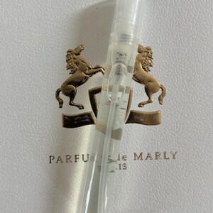 May include: A clear perfume sample vial with a spray nozzle, resting on a white surface with a gold emblem featuring two rearing horses. The text "PARFUMS de MARLY" is visible.