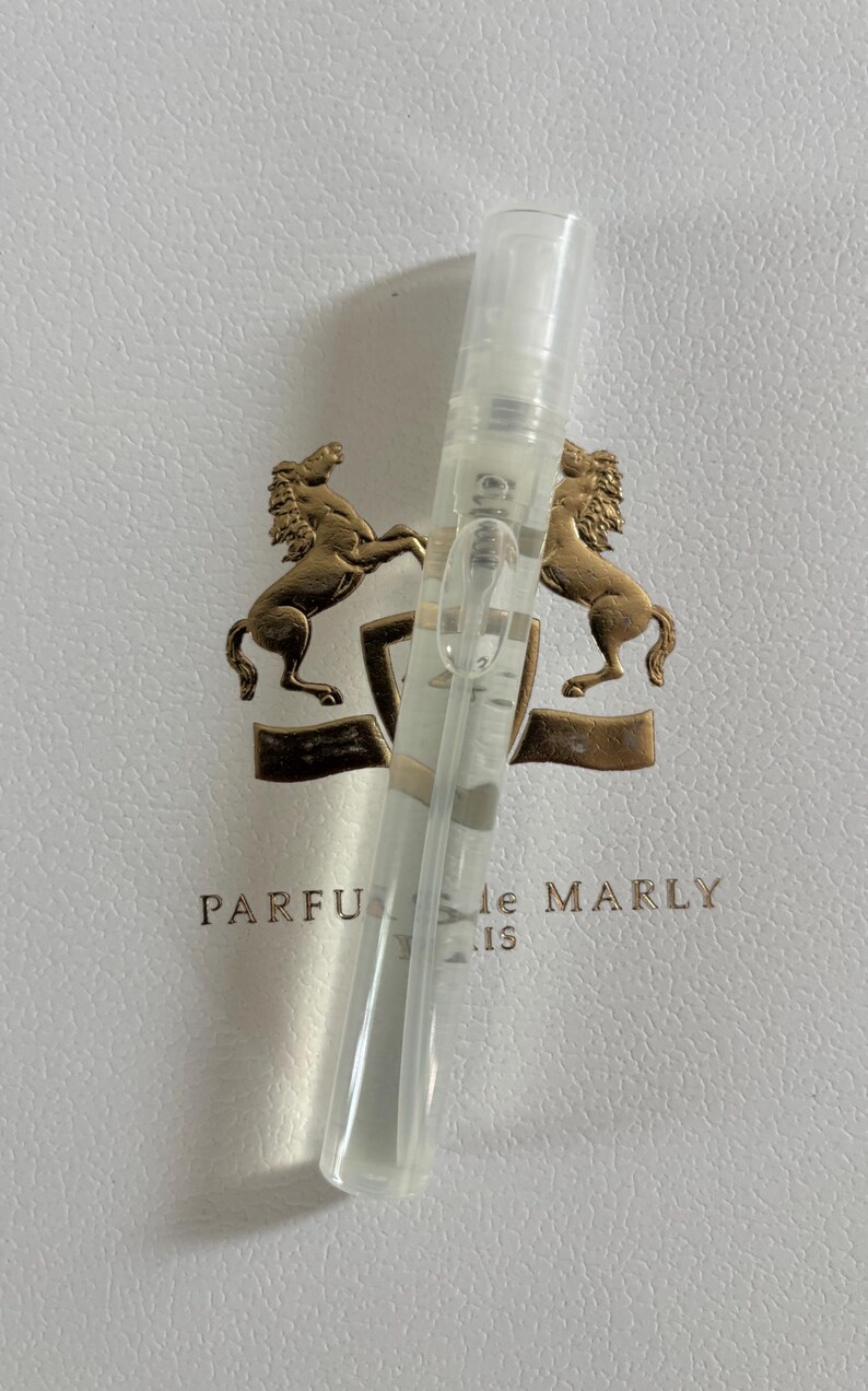May include: A clear perfume sample vial with a spray nozzle, resting on a white surface with a gold emblem featuring two rearing horses. The text "PARFUMS de MARLY" is visible.