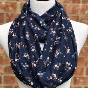 May include: A navy blue infinity scarf featuring a repeating pattern of small white, peach, and red flowers. The scarf is draped in a circular loop, showcasing the fabric's soft texture and the dense, evenly distributed floral print across the surface.