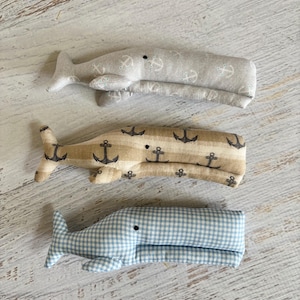 May include: Three fabric whale plush toys. The top whale is light gray with white anchor patterns. The middle whale is tan with black anchor patterns. The bottom whale is blue and white gingham.
