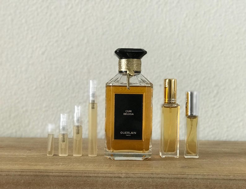 May include: A collection of perfume bottles, including a large bottle of Guerlain Cuir Beluga perfume. The bottles are arranged in a row on a wooden surface.