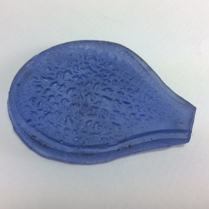Puede incluir: Un plato de vidrio azul claro con una superficie texturizada. El plato tiene forma de lágrima y tiene un borde ligeramente elevado.