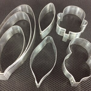 Könnte beinhalten: Satz mit sechs Metall-Ausstechformen in verschiedenen Formen, darunter Blätter, Blumen und eine Glocke. Die Ausstechformen sind silberfarben und haben eine glänzende Oberfläche.