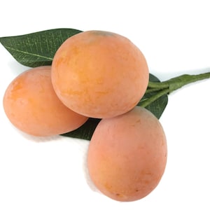 May include: Three artificial peach-colored fruits arranged on a green stem with two dark green leaves. The fruits have a smooth, matte texture and a rounded, slightly oval shape. The stem is wrapped in green material, extending from the fruit cluster.