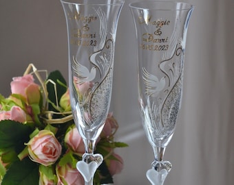 May include: Two clear glass champagne flutes with gold lettering and a white dove design. The flutes have a heart-shaped base and are personalized with the names "Maggie & Danni" and the date "16.05.2023".