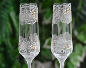 May include: Two clear glass champagne flutes with white floral designs and gold accents. The flutes are on a reflective surface.
