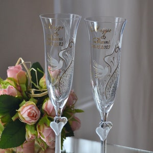 May include: Two clear glass champagne flutes with gold lettering and a white dove design. The flutes have a heart-shaped base and are personalised with the names "Maggie & Danni" and the date "16.05.2023".