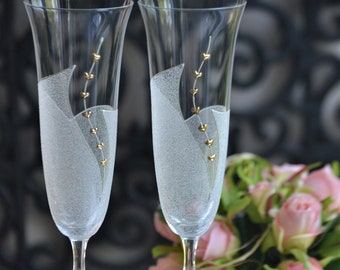 May include: Two clear glass champagne flutes with gold accents and a frosted design. The flutes are on a reflective surface with a bouquet of pink roses in the background.