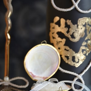 May include: A white seashell ring holder with a gold trim and a white ribbon. The ring holder is sitting on a black surface with a gold and black bottle in the background.