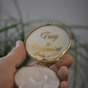 May include: A white ring box with a gold trim and a white satin ribbon. The lid is open and has the inscription "Troy & Francine July 8th" in gold lettering.