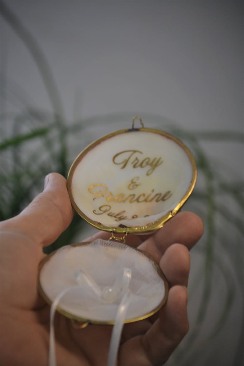 May include: A white ring box with a gold trim and a white satin ribbon. The lid is open and has the inscription "Troy & Francine July 8th" in gold lettering.