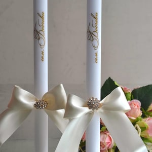 Può includere: Due candele cilindriche bianche alte, ciascuna con un'iscrizione dorata in corsivo e un nastro di raso color crema annodato a fiocco. Un ornamento circolare in strass trasparente è centrato su ogni fiocco, aggiungendo un tocco di luce.