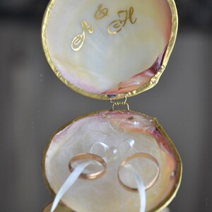 May include: A white seashell ring box with a gold trim and the initials "J & H" inscribed on the lid. Two gold wedding bands are inside the box, tied with a white ribbon.