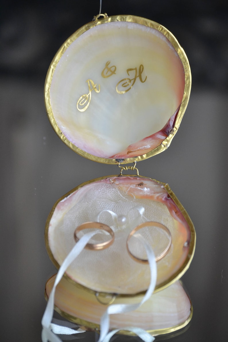 May include: A white seashell ring box with a gold trim and the initials "J & H" inscribed on the lid. Two gold wedding bands are inside the box, tied with a white ribbon.