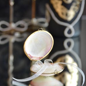 May include: A small, white seashell ring box with a gold trim. The box is open and has a white tulle bow inside. The box is sitting on a black surface.