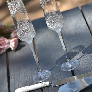 May include: Two clear glass champagne flutes with white floral designs and a silver cake server with a white handle with a floral design.