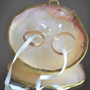 May include: Two gold wedding bands are displayed inside a seashell ring bearer pillow. The pillow is made of a white fabric with a delicate lace pattern. The bands are tied together with a white satin ribbon.