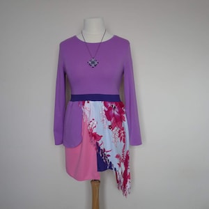 Puede incluir: Un vestido morado de manga larga con una falda de estampado floral rosa y blanco. El vestido tiene un cinturón morado y un collar con un colgante geométrico.