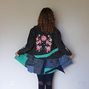 May include: A black denim jacket with floral embroidery on the back. The jacket is worn over a teal and blue denim skirt. The person is wearing black tights with floral designs.