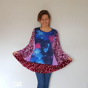 Pode incluir: Top túnica de manga comprida com estampa de galáxia no painel frontal, mangas magenta e roxas. A bainha inferior é de tecido bordô com padrões de estrelas brancas. A modelo usa jeans azuis e meias pretas.