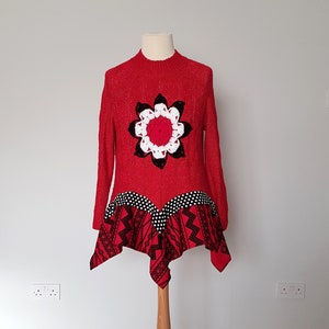 Könnte beinhalten: Ein roter Pullover mit einem gehäkelten Blumendesign in Schwarz, Weiß und Rot. Der untere Teil des Pullovers hat einen gemusterten Stoff mit schwarzen und weißen Punkten und geometrischen Mustern. Auf einer Schaufensterpuppe präsentiert.
