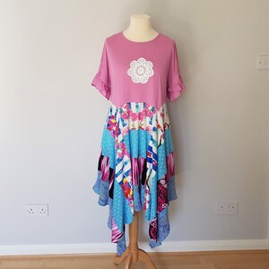May include: A pink dress with a white crocheted flower on the bodice. The skirt is made of a patchwork of colorful fabrics with floral and geometric patterns.