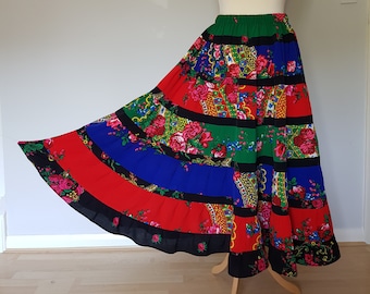 Op de afbeelding: Een lange, gelaagde rok met een levendig bloemenpatroon en een mix van kleuren, waaronder rood, blauw, groen en zwart. De rok wordt op een mannequin gepresenteerd, waarbij het vloeiende ontwerp en de gedetailleerde print worden benadrukt. De zoom van de rok is afgewerkt met een zwarte bies.