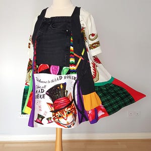 Puede incluir: Un vestido único de estilo patchwork con un peto de mezclilla negro y una base de camiseta blanca. El vestido presenta paneles de tela coloridos, incluido un tartán verde, y un gráfico del Gato de Cheshire con el texto "Welcome to the MAD HOUSE We all MAD HERE."