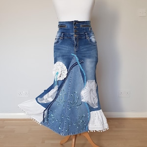 Op de afbeelding: Een lange spijkerbroekrok met hoge taille en decoratieve elementen. De rok heeft witte kant en geborduurde bloemendetails. De rok wordt op een mannequin gepresenteerd. De rok heeft een blauw-wit kleurenschema.