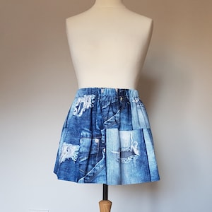 May include: A blue denim skirt with a patchwork design, displayed on a mannequin. The skirt features various shades of blue denim with distressed details, including rips and visible stitching. The skirt has an elasticated waistband.
