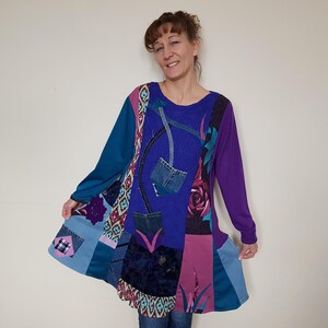 Könnte beinhalten: Eine Patchwork-Tunika in Blau-, Lila- und Rosatönen. Das langärmlige Oberteil hat Jeans-Taschen und florale Akzente. Die Tunika wird von einer Person getragen.