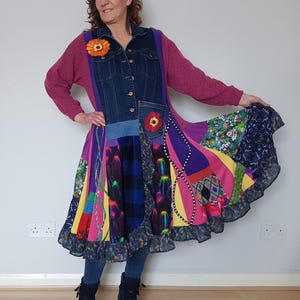 Könnte beinhalten: Ein farbenfrohes Patchwork-Kleid mit einer Jeansjacke. Das Kleid weist eine Vielzahl von Mustern und Farben auf, darunter Lila, Rosa, Gelb und Blau. Eine Blumenbrosche ziert die Jacke. Die Person trägt eine blaue Jeans und schwarze Stiefel.
