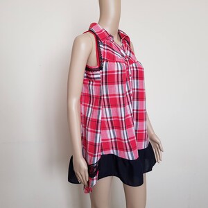 May include: A red and white plaid sleeveless shirt with a black collar and a black ruffle trim. The shirt has a button-up front.