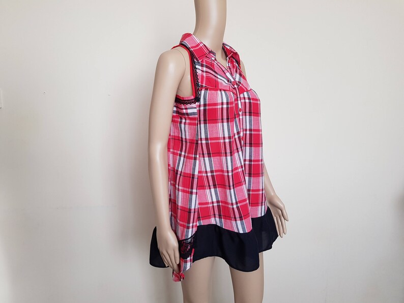 May include: A red and white plaid sleeveless shirt with a black collar and a black ruffle trim. The shirt has a button-up front.