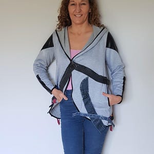 May include: A grey hooded jacket with black faux-leather panels on the shoulders and sleeves. The jacket has denim patches and a belt detail. The person is wearing blue jeans and a pink top.