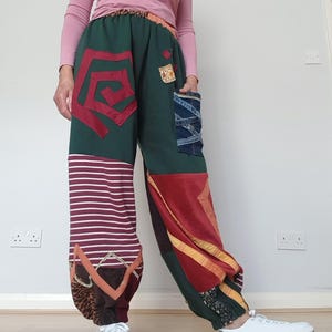 May include: Loose-fitting patchwork trousers featuring a dark green base with various fabric panels including maroon, white horizontal stripes, denim, and animal print. The design includes a large red spiral appliqué, a denim pocket, and elasticated cuffs.