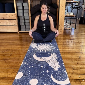 Peut inclure: Un tapis de yoga bleu marine avec un motif c&eacute;leste repr&eacute;sentant des soleils, des lunes, des nuages et des &eacute;toiles. Le tapis est rectangulaire et a une bordure rose clair. La personne porte un d&eacute;bardeur noir avec un motif de phases lunaires.