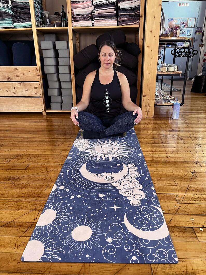 Peut inclure: Un tapis de yoga bleu marine avec un motif c&eacute;leste repr&eacute;sentant des soleils, des lunes, des nuages et des &eacute;toiles. Le tapis est rectangulaire et a une bordure rose clair. La personne porte un d&eacute;bardeur noir avec un motif de phases lunaires.
