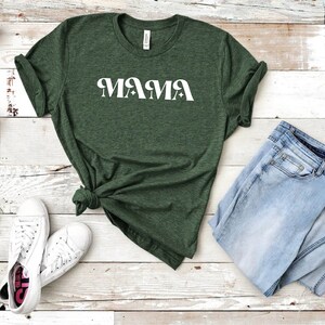 May include: A green t-shirt with the word "MAMA" printed in white on the front. The shirt is laid out on a white wooden surface with a pair of white sneakers and a pair of blue jeans.