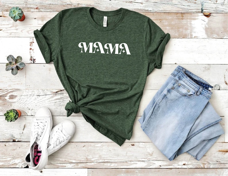 May include: A green t-shirt with the word "MAMA" printed in white on the front. The shirt is laid out on a white wooden surface with a pair of white sneakers and a pair of blue jeans.