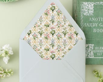May include: A light blue envelope with a floral patterned interior, alongside a green plate, a vintage book titled "Another Hardy Garden Book", and two "Larkspur" seed packets. White flowers and a light green background complete the scene.