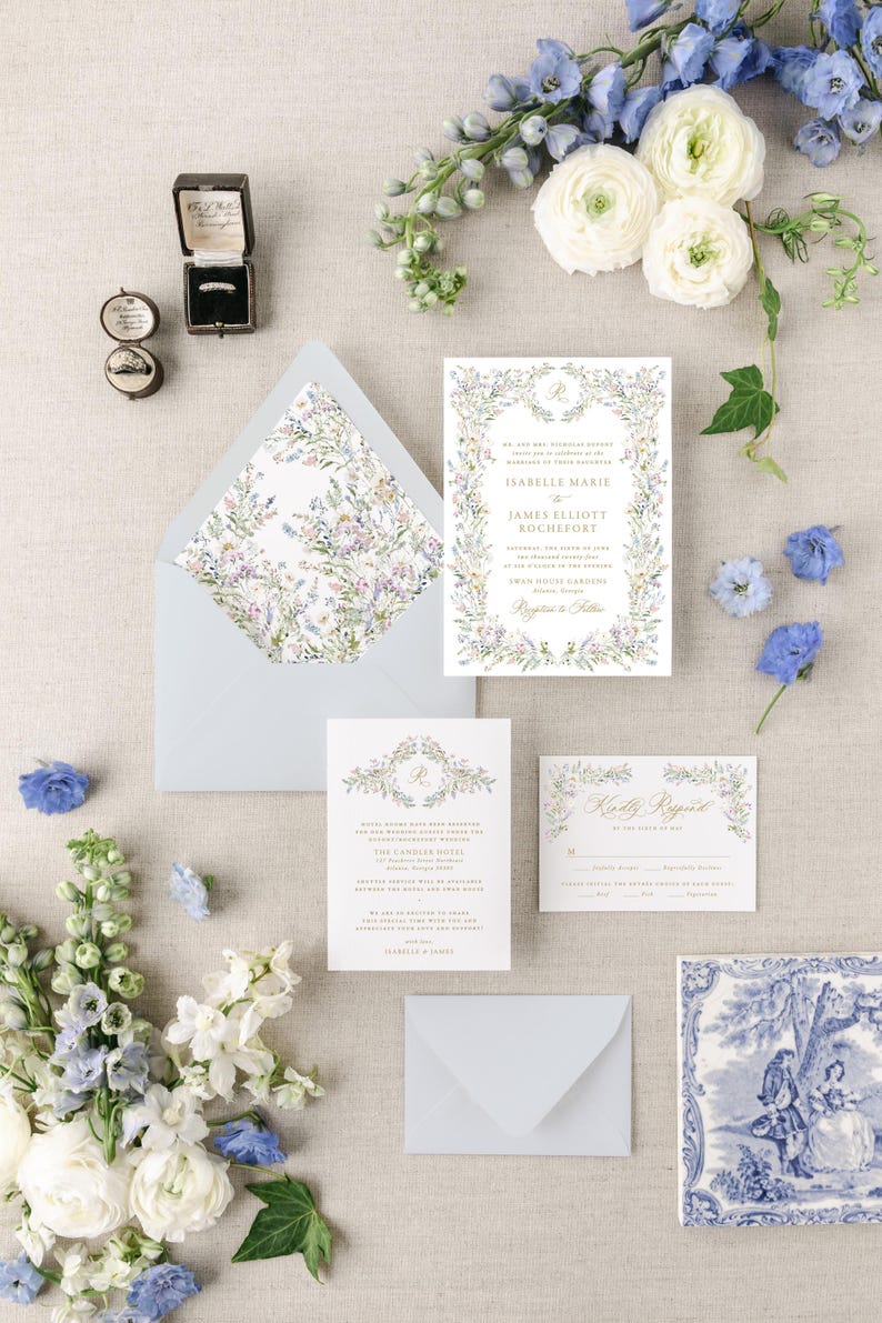 May include: A wedding invitation suite with a floral design. The invitation features a wreath of watercolor flowers and greenery with the couple's names and wedding details. The RSVP card features a similar floral design with a gold script font. The envelope liner features a floral pattern.