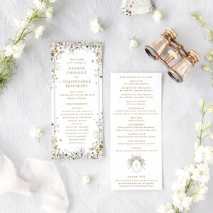 a wedding invitation with a floral design, a pair of binoculars, and some white flowers arranged on a white background.