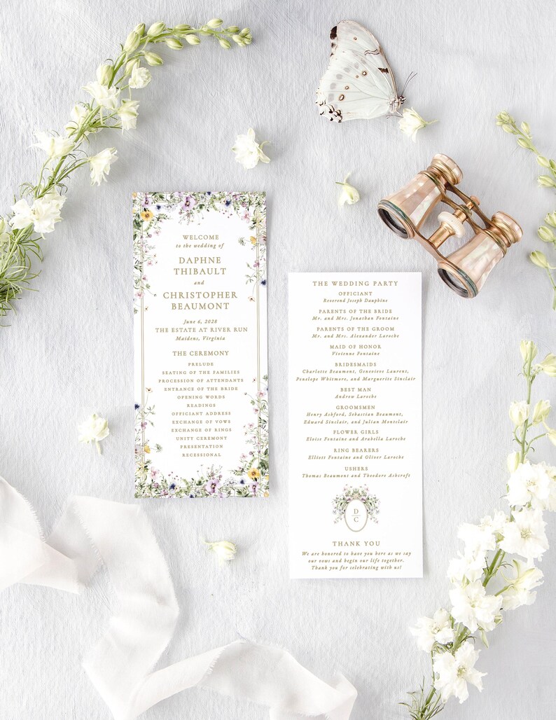 a wedding invitation with a floral design, a pair of binoculars, and some white flowers arranged on a white background.