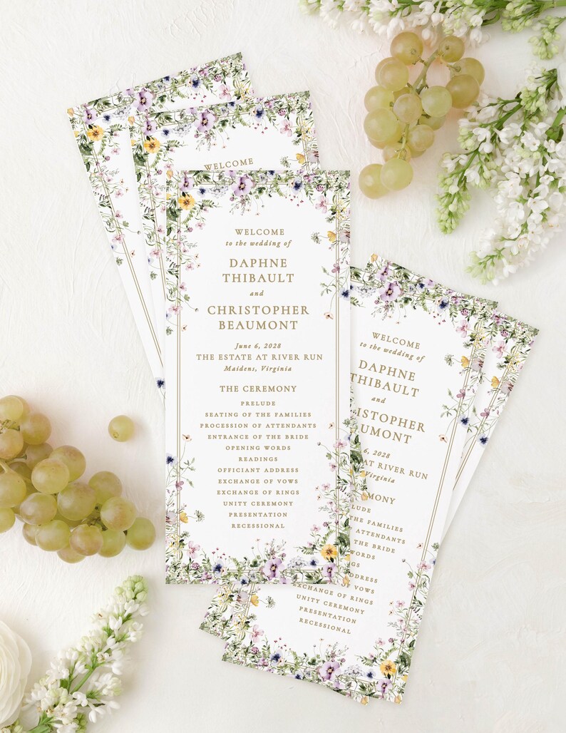 a set of wedding invitations with a floral design, placed on a white background alongside some grapes and flowers.