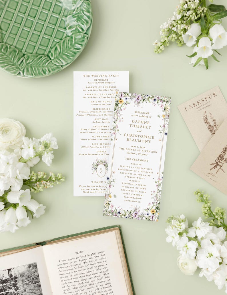 a wedding invitation with a floral design, surrounded by various wedding-related items such as a green plate, white flowers, and a book.