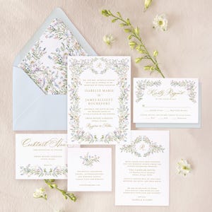 May include: A floral wedding invitation suite with a blue envelope, a white invitation card with a floral border and gold lettering, a white RSVP card with a floral border, and two white cards with floral borders and details about the cocktail hour and hotel accommodations.