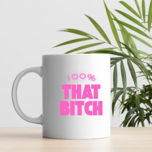 May include: A white ceramic mug featuring the text 100% THAT BITCH printed in a bold, bright pink font. The mug has a standard cylindrical shape with a curved handle on the left side, designed for holding hot or cold beverages in a simple style.