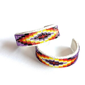 May include: Two beaded bracelets with a geometric pattern in shades of purple, yellow, orange, red, and black. The bracelets are made of leather and have a white background.
